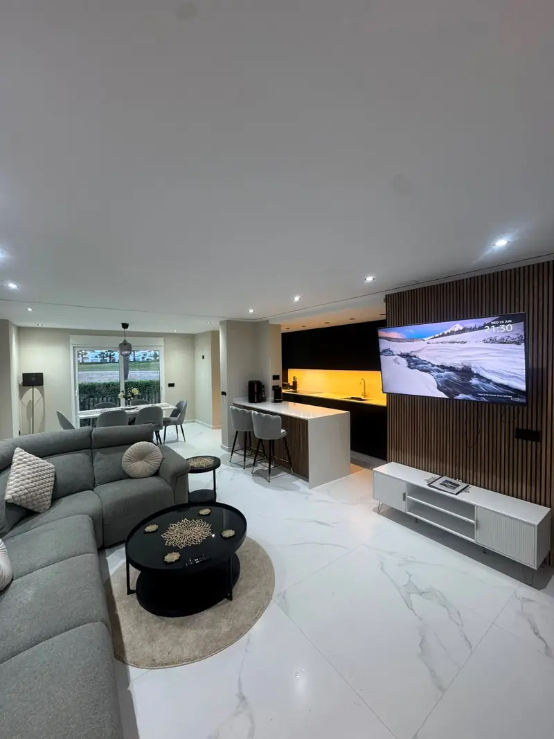 Open-plan ground floor with grey corner sofa, wall-mounted TV on walnut slat feature wall, marble floors and dining area with garden view
