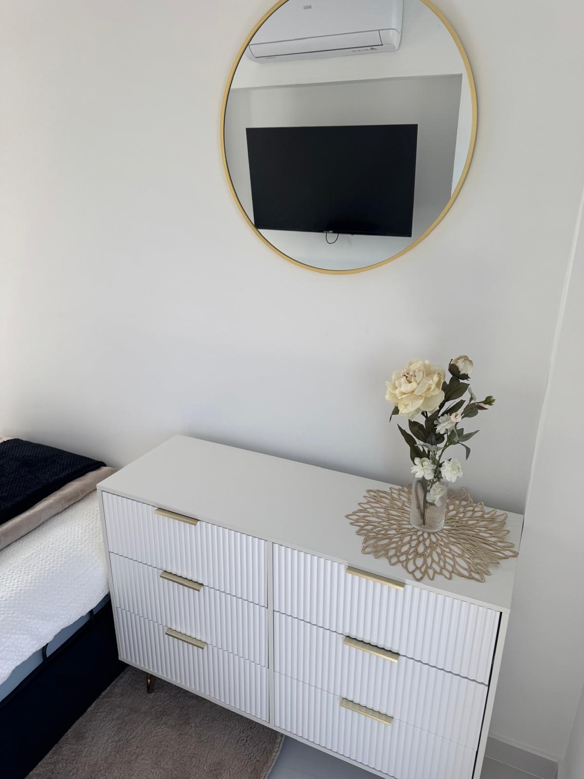 White fluted six-drawer dresser with brass handles beneath a round gold-framed mirror