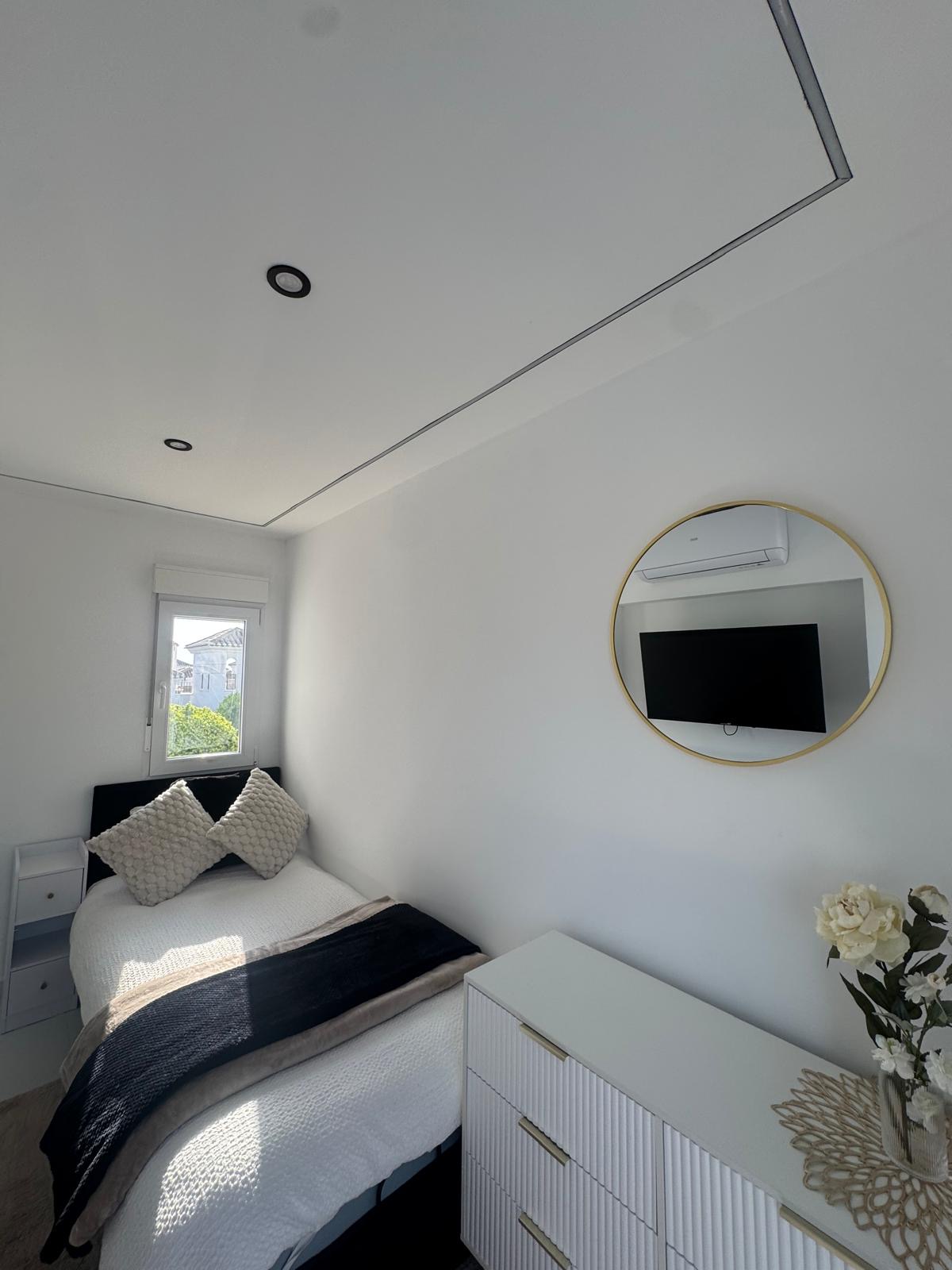 Third bedroom with single bed, gold-framed round mirror and fluted white dresser in bright natural light